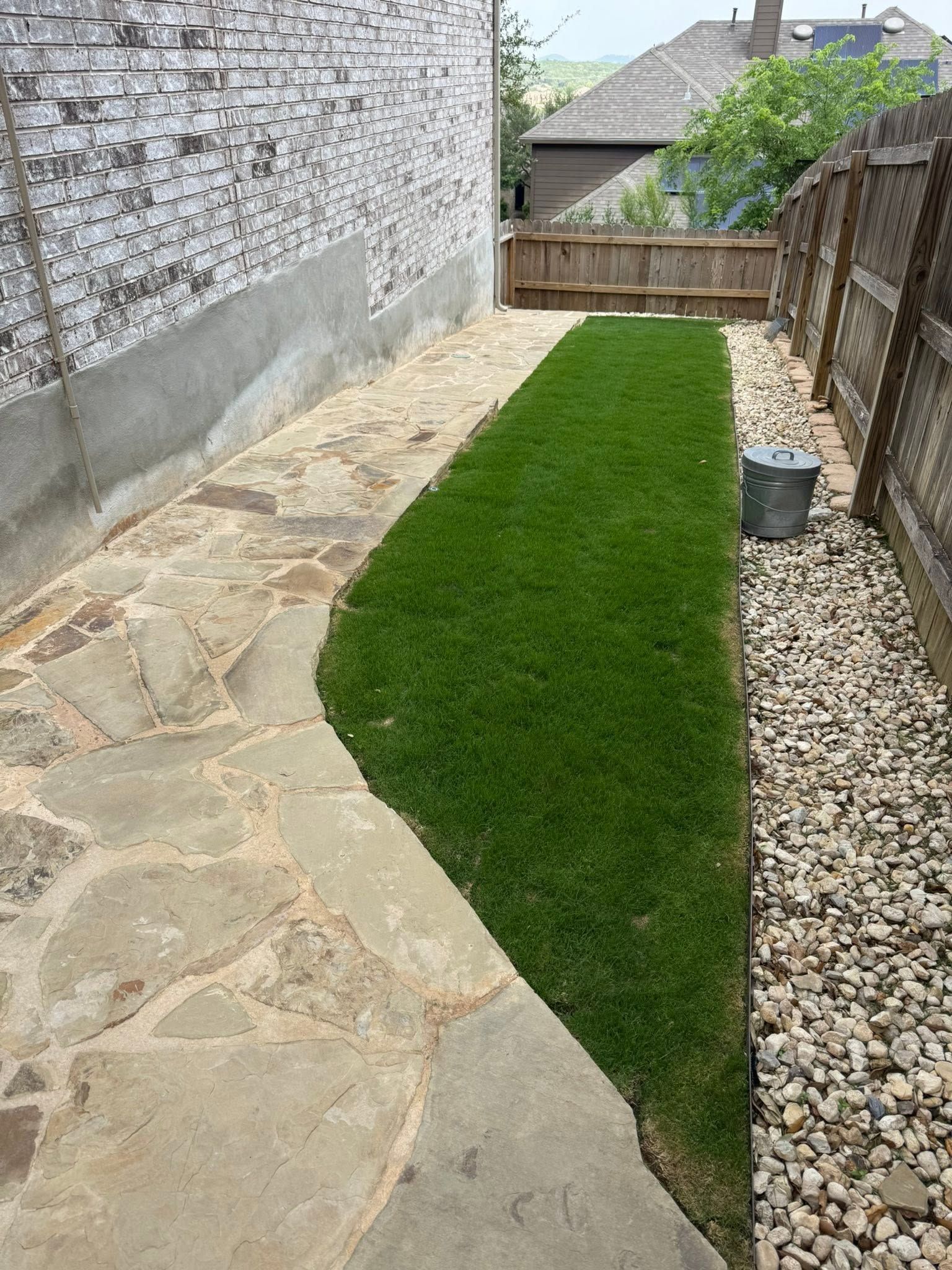 Narrow backyard with stone walkway, strip of green grass, gravel border, and wooden fence