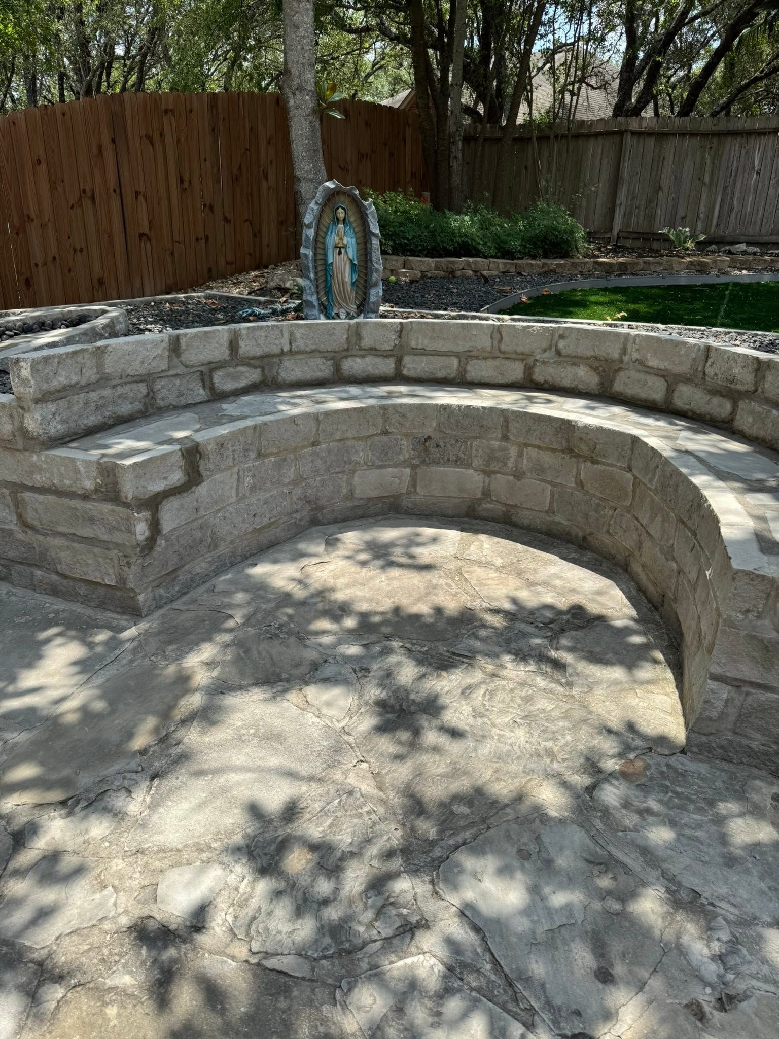 Curved stone bench around a fire pit in a backyard patio, with trees and a wooden fence behind it