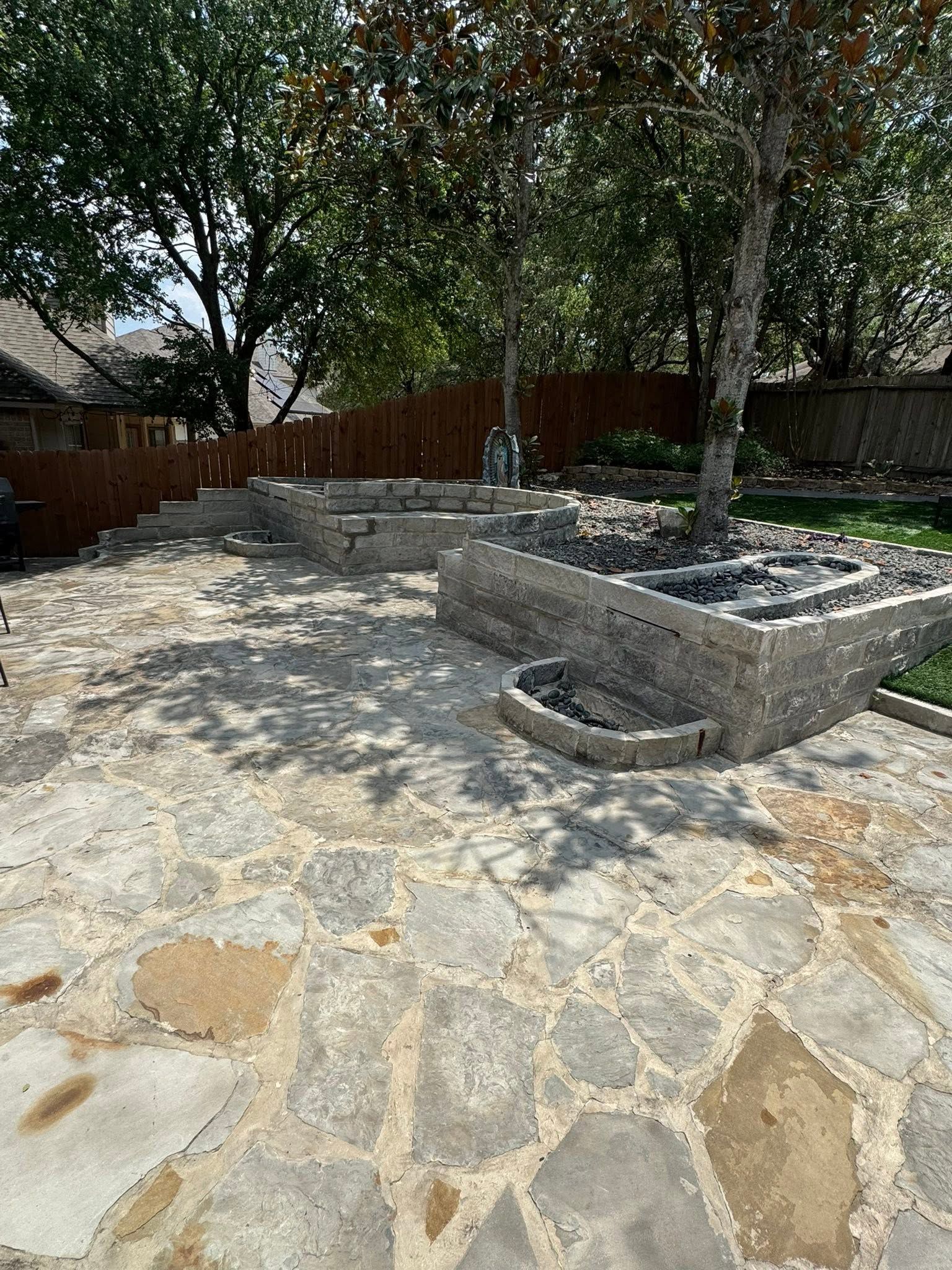 Stone patio with raised garden beds and mature trees in a backyard.