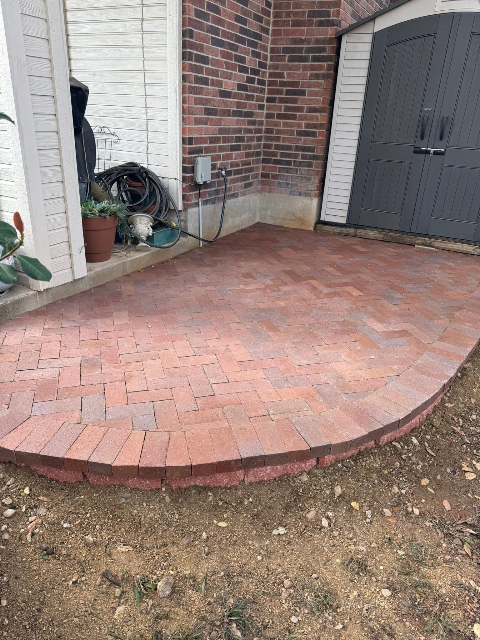 Curved brick patio beside a white wall and dark door, with a small planter and hose reel.
