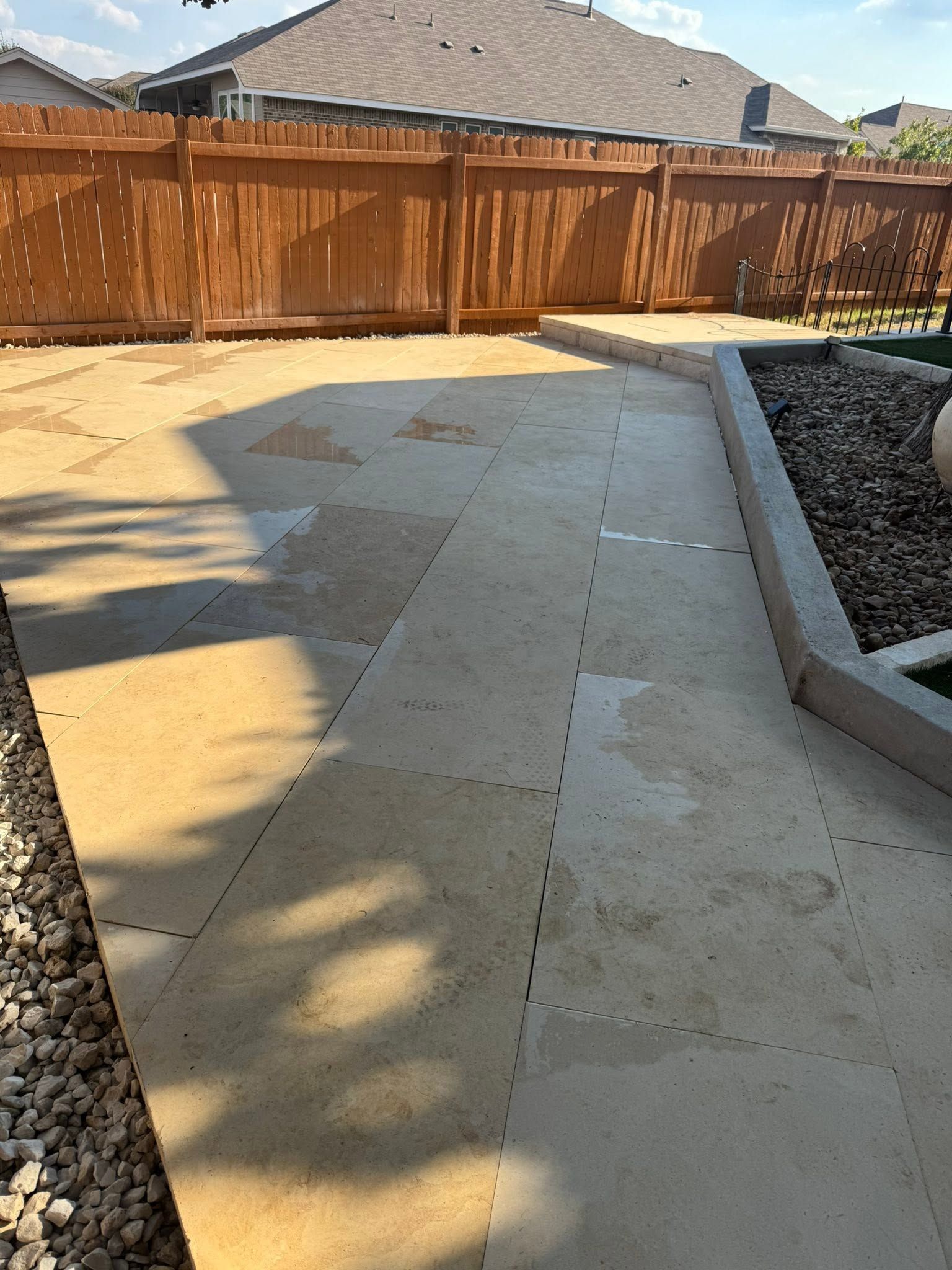 Concrete backyard patio beside a wooden fence, bordered by rock landscaping and a raised planter bed.