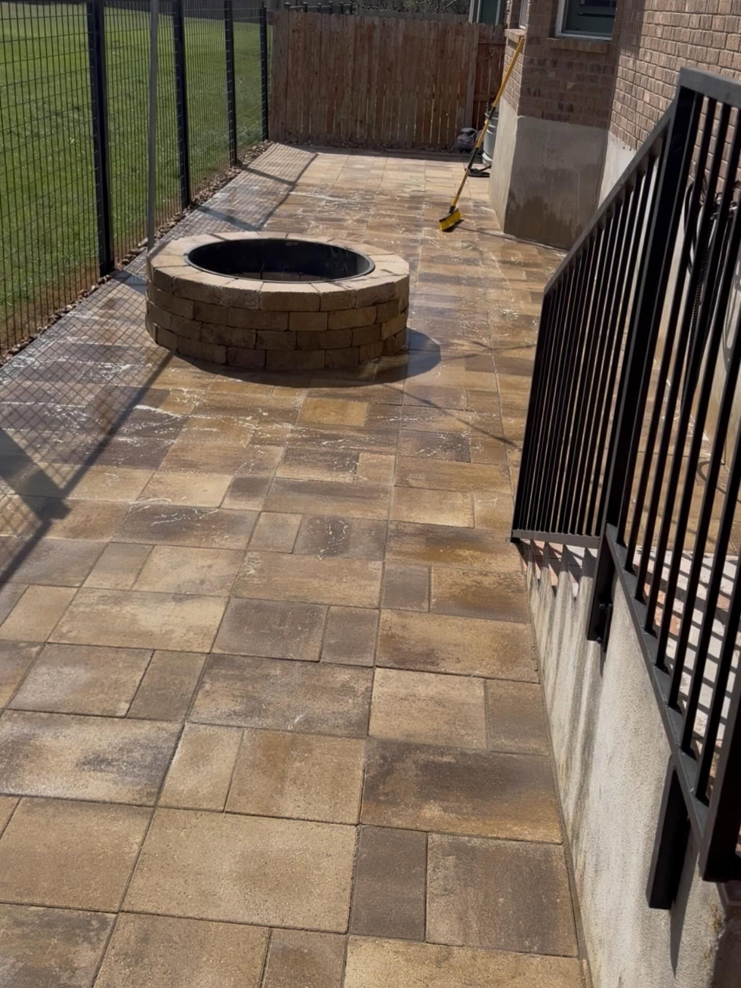 Stone patio with a circular fire pit, black fence, and gate in a fenced backyard.