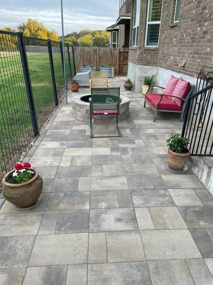 Backyard patio with gray pavers, black fence, brick house, and patio furniture with potted plants.