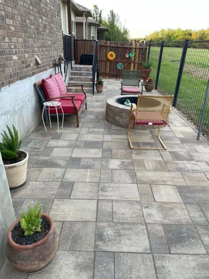 Patio with gray pavers, red bench, fire pit, potted plants, and wicker chair beside a house and fence.