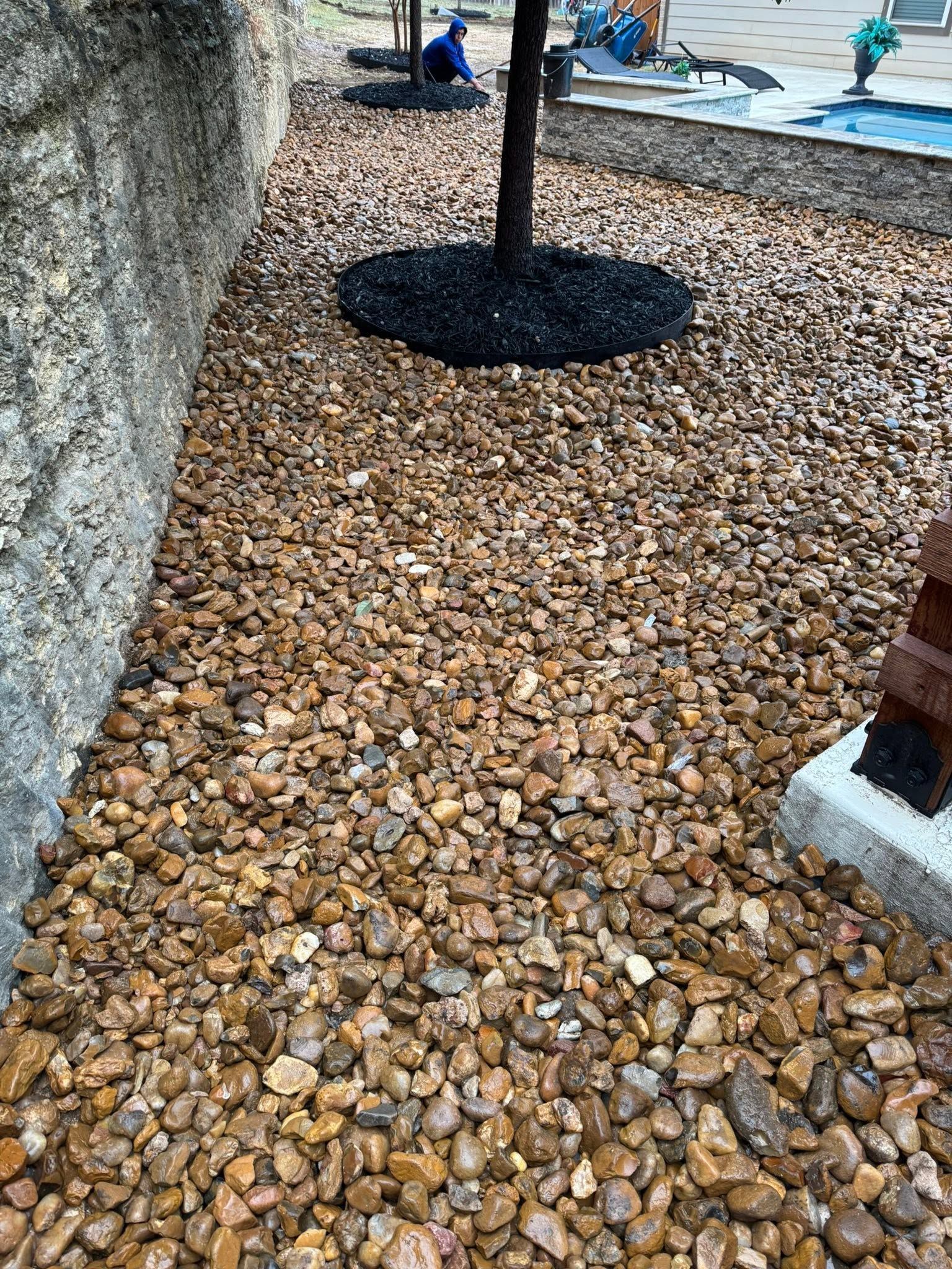 Pebble ground cover around a tree, with a large rock on the left and a paved area in the background