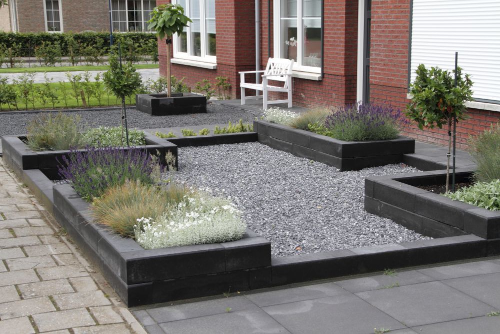 Raised black planters with gravel and shrubs in a small front garden beside a brick building.