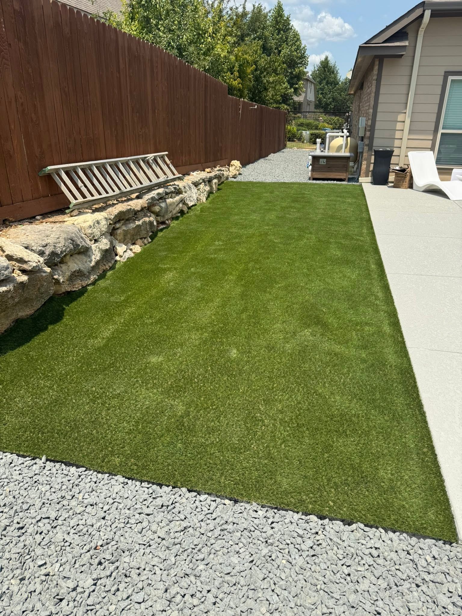 Backyard with artificial grass, gravel path, wooden fence, and a house along the right side
