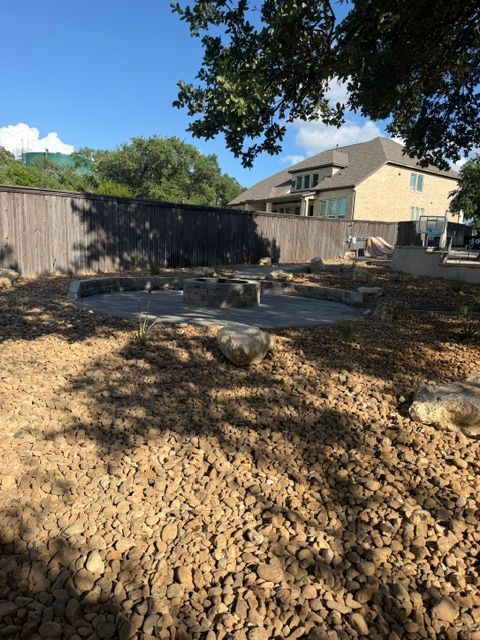 Backyard with a cracked dirt surface, a stone patio, wooden fence, and a house in the background.