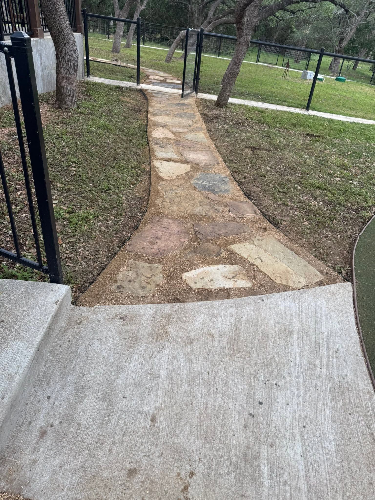 Concrete ramp leading to a narrow dirt path beside a black metal fence in a grassy area.