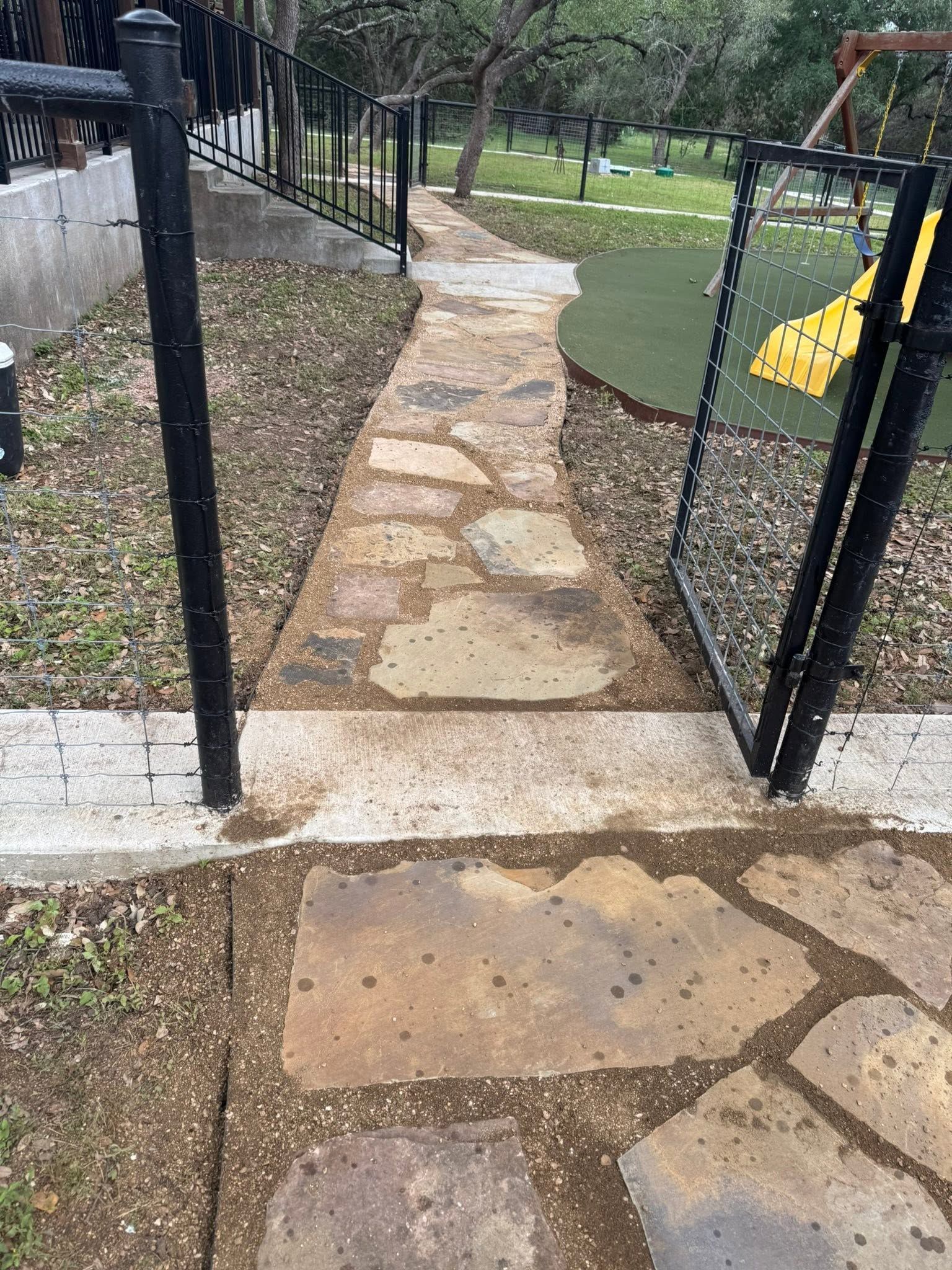 Stone path through a park entrance, flanked by black fences, leading to a grassy play area with a yellow slide.