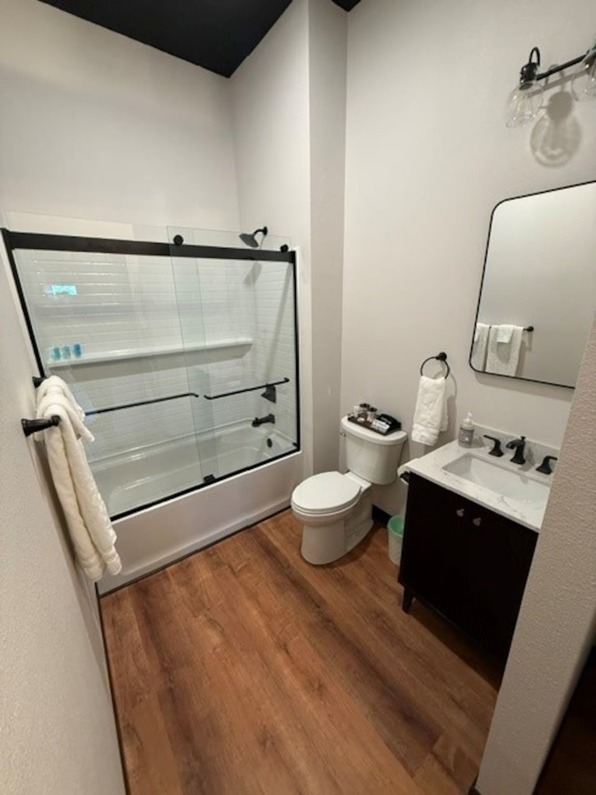Oelwein, Iowa modern bathroom with shower-tub, toilet, vanity sink, mirror, and wood-look flooring.