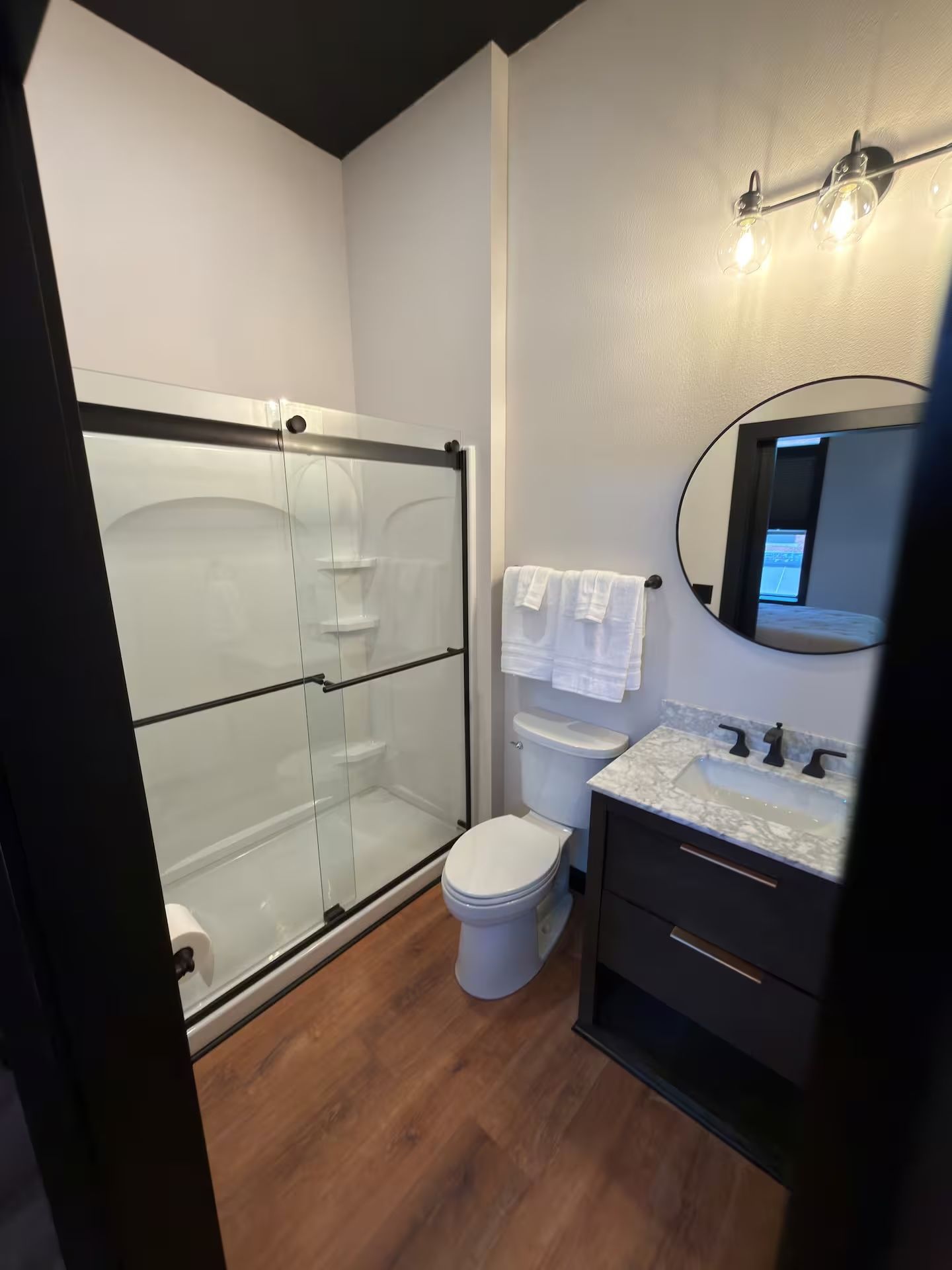 Oelwein, Iowa compact modern bathroom with glass shower, toilet, vanity, round mirror, and wood-look floor.