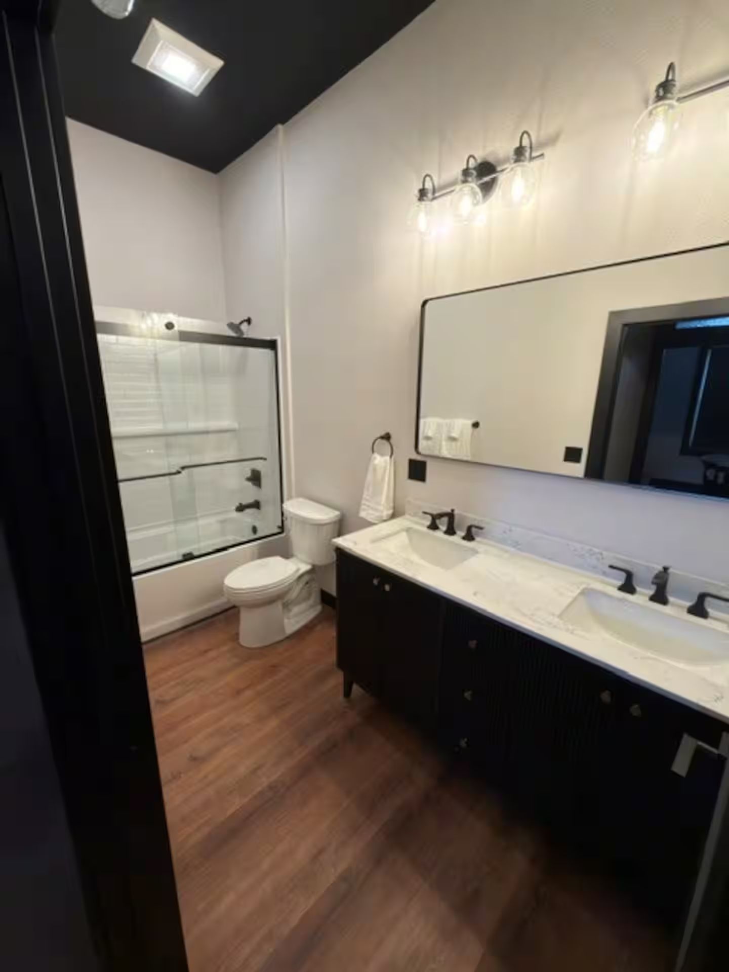 Oelwein, Iowa modern bathroom with bathtub, toilet, vanity, large mirror, white walls, and wood-look floor.
