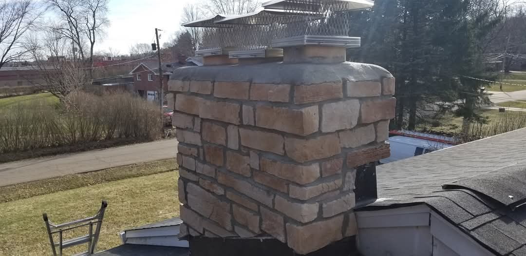 Brick chimney with metal chimney caps on a shingled roof. Houses and trees in the background.