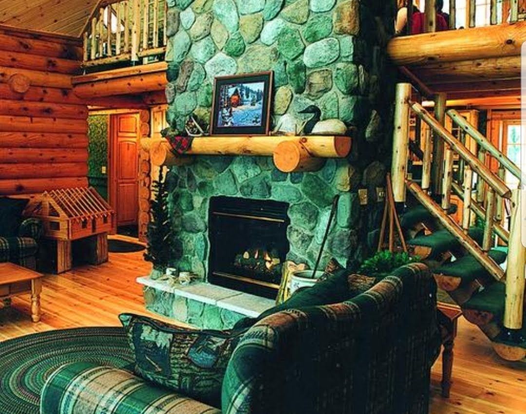 Cozy cabin interior with a stone fireplace, log walls, wooden stairs, and a plaid armchair.
