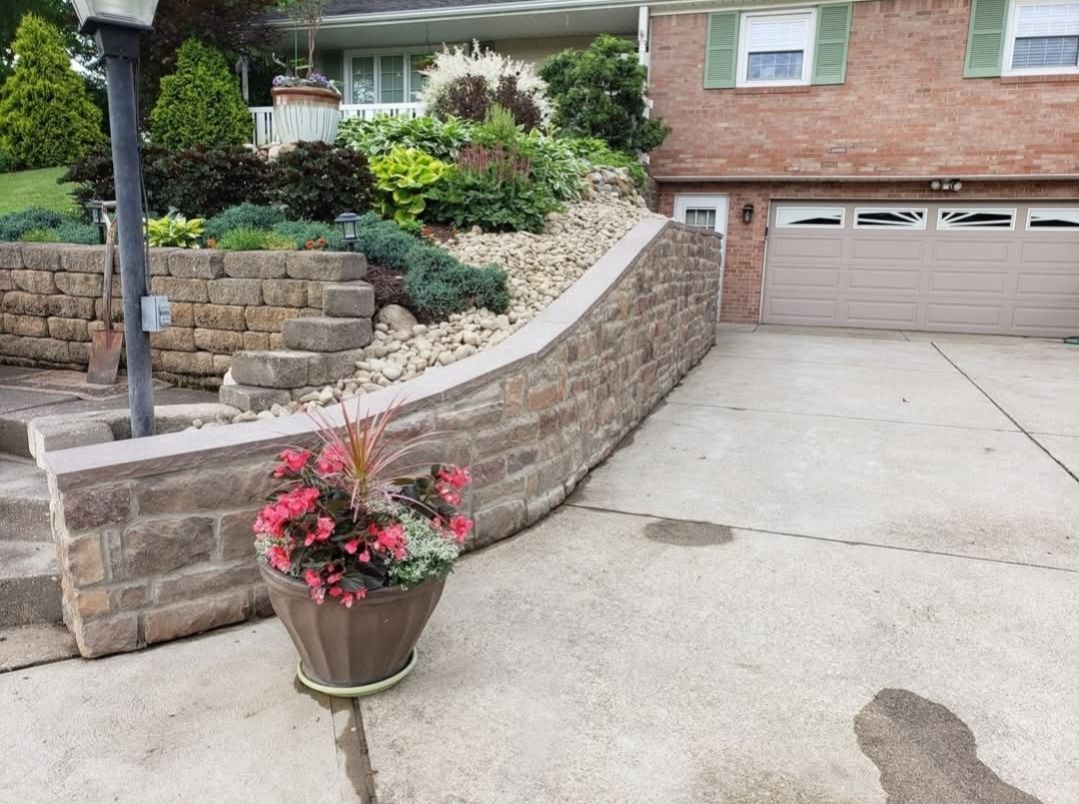 Stone retaining wall with steps, a house, and a garage. A planter with pink flowers sits in front.