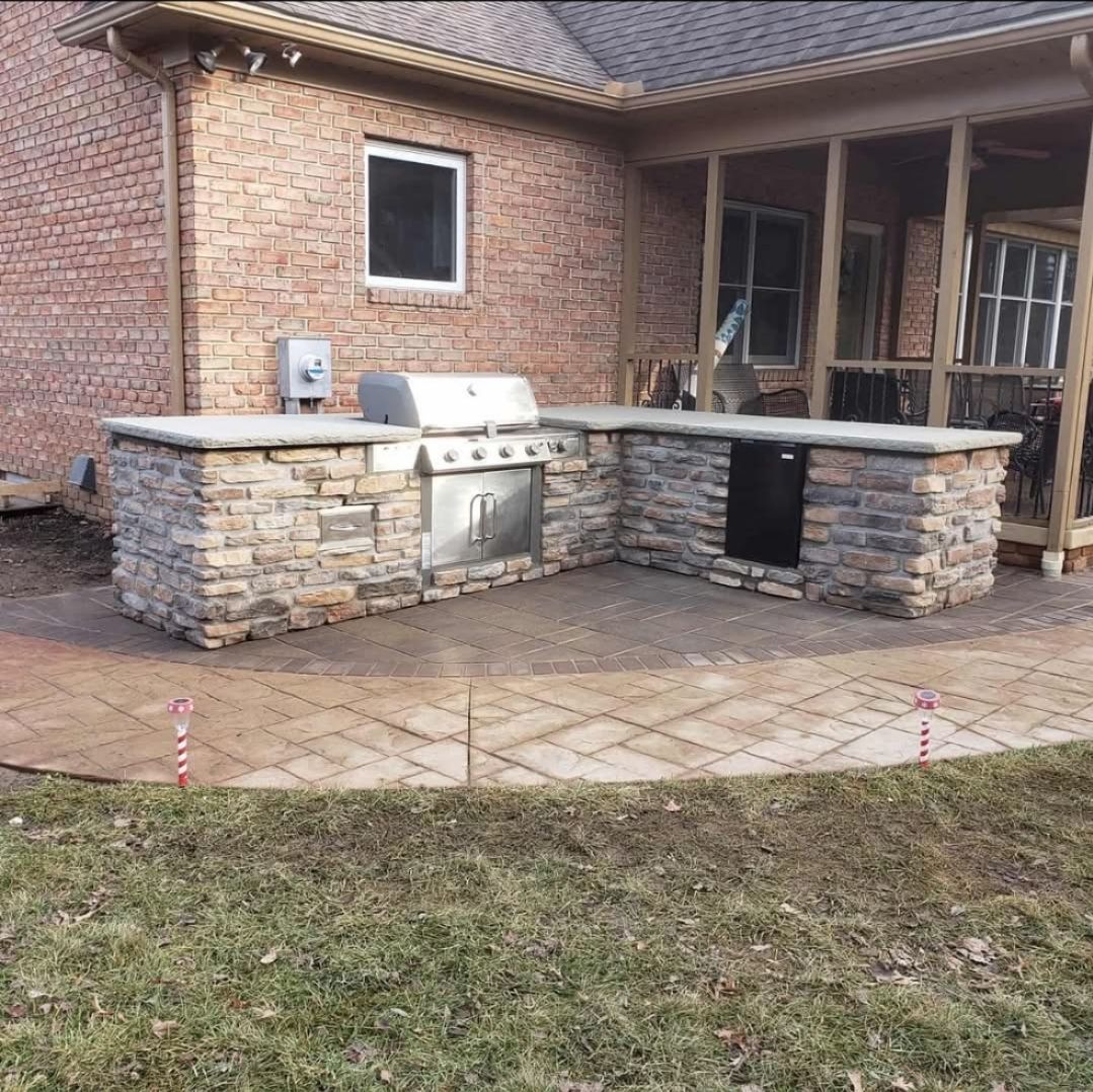 Outdoor stone built-in grill and counter with brick house background and patterned patio.