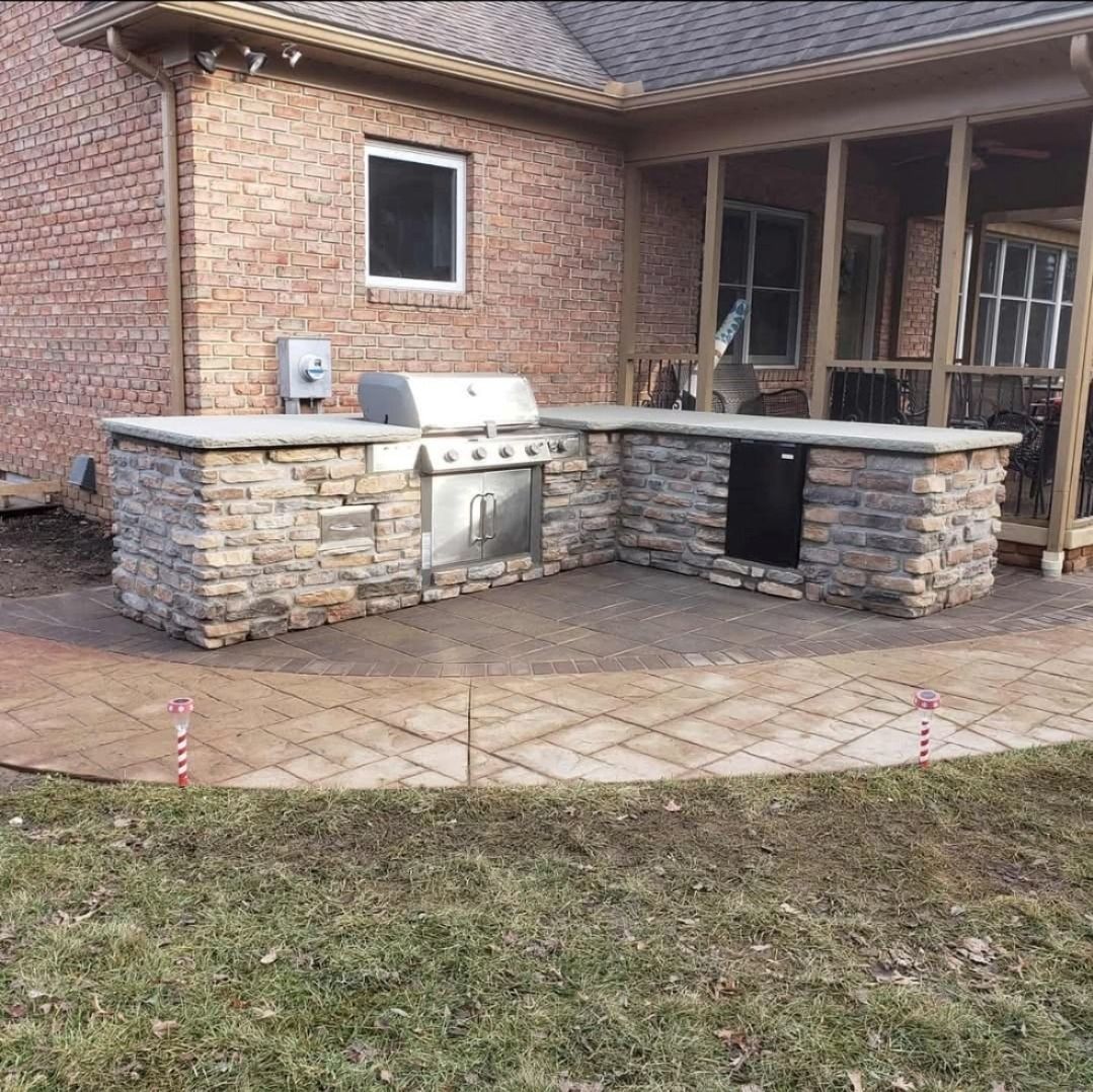 Outdoor stone kitchen with grill, counter space, and a small refrigerator, set on a patio near a brick house.