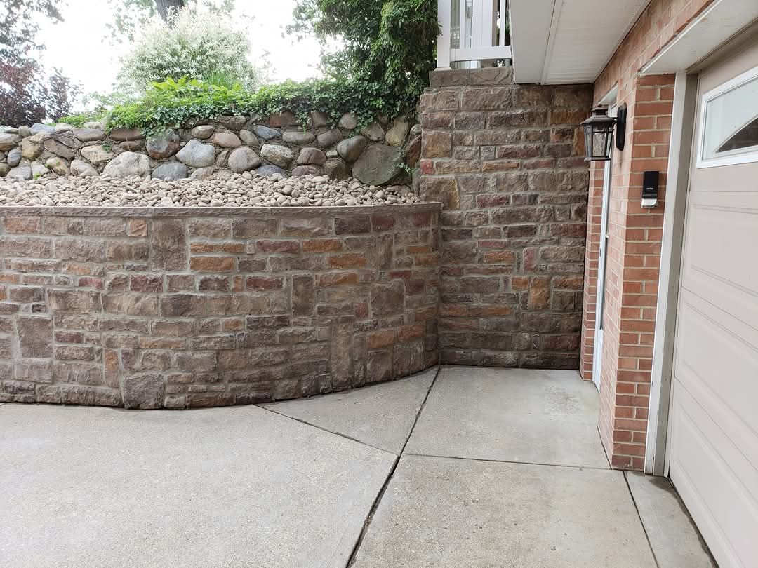 Brick retaining wall beside a garage, with a concrete driveway in the foreground and greenery above the wall.