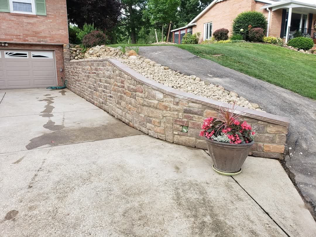 Brick retaining wall next to a driveway, with a potted plant in the foreground and a grassy hill in the background.