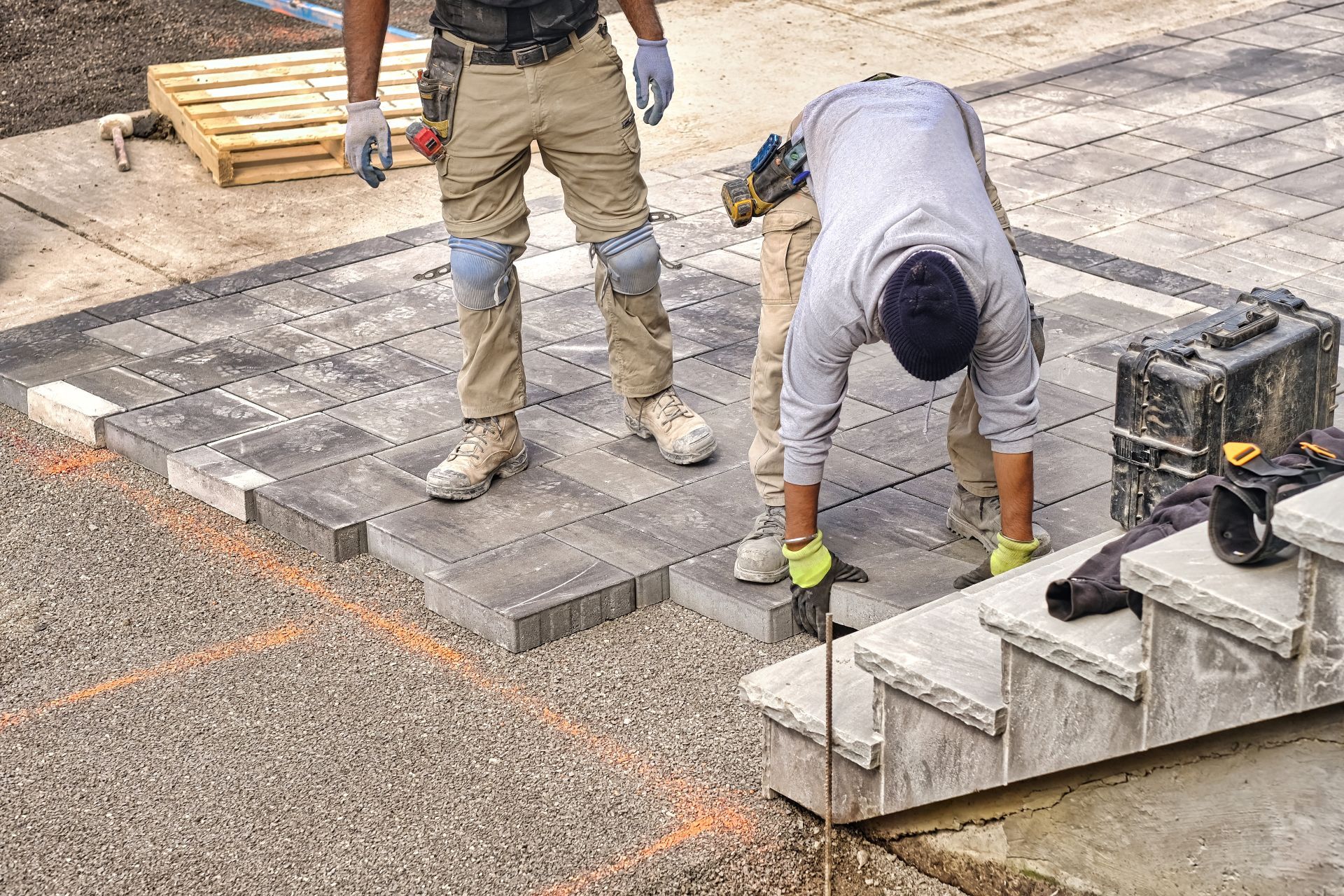 Two workers laying stone pavers on a patio with a gravel base near concrete steps.