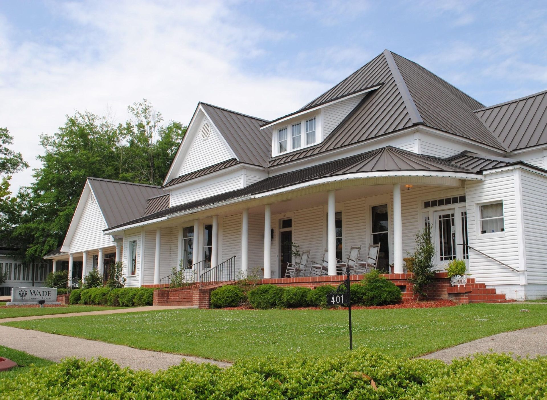 White farmhouse with wraparound porch, gray roof, and red brick foundation on a green lawn.