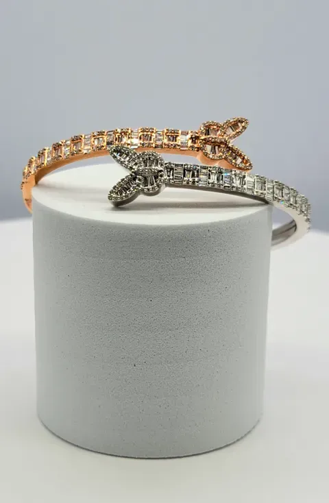 Two jeweled bracelets, one gold and one silver, displayed on a white cylindrical stand.