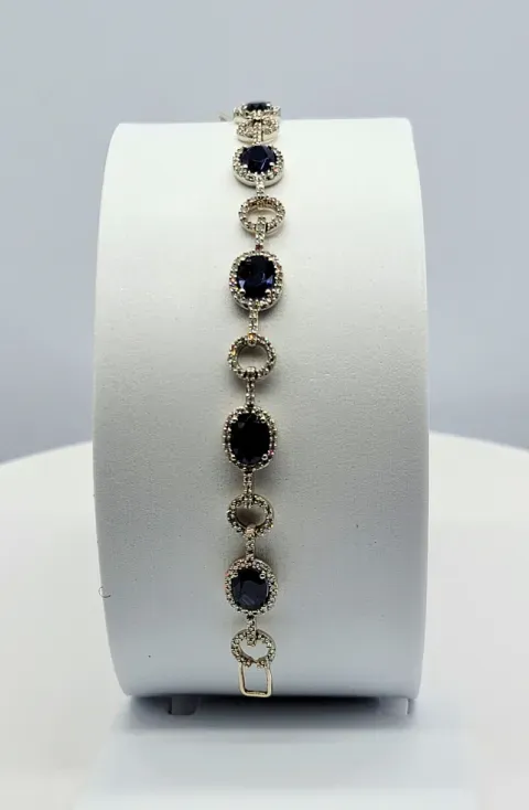 Gold bracelet with oval dark blue and clear gemstones displayed on a white stand