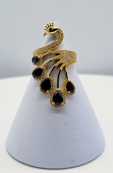 Gold peacock-shaped ring with black stones displayed on a white cone stand.