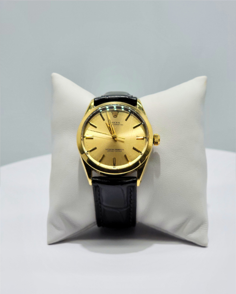 Gold wristwatch with black leather strap displayed on a white pillow against a light gray background