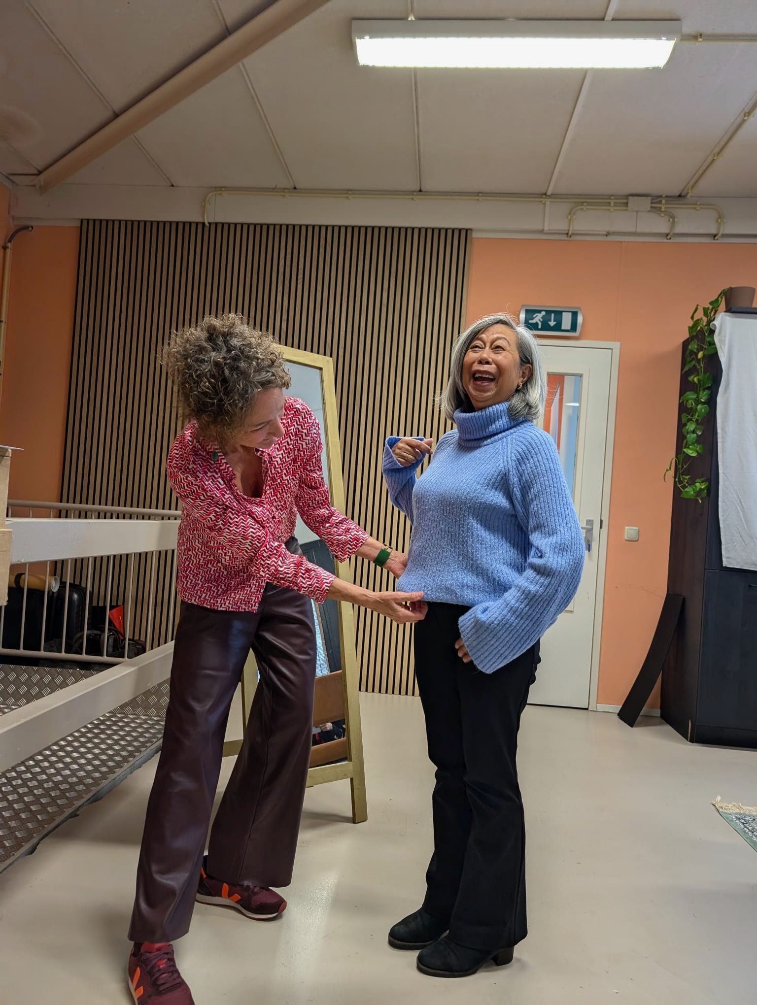 Twee mensen in een studio; de ene maakt de zoom van een blauwe gebreide trui van de ander recht, die lacht.