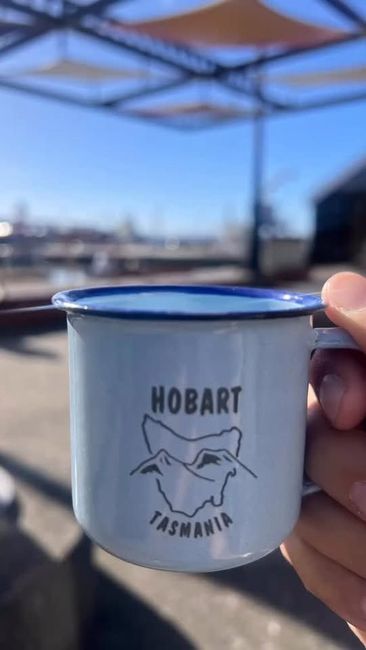 Hand holding a white Hobart Tasmania enamel cup against a blue sky outdoors