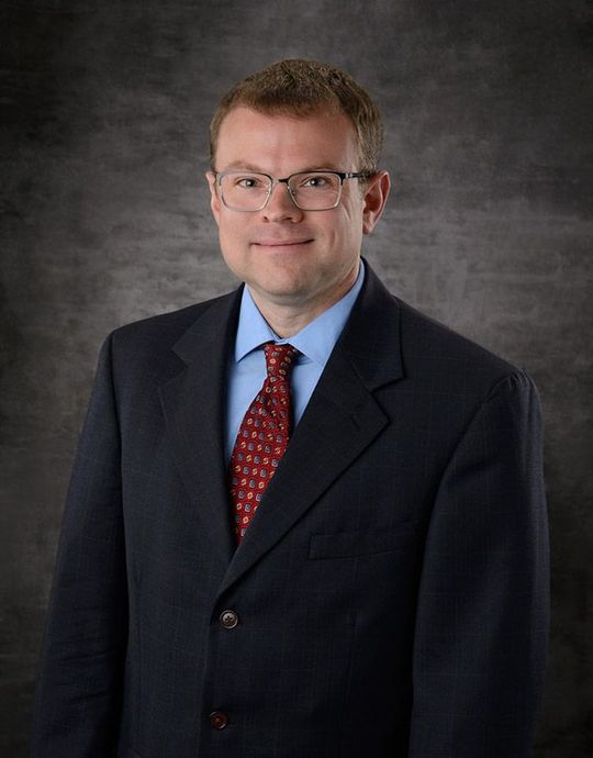 Man in a dark suit, blue shirt, and red tie, smiling against a gray studio backdrop