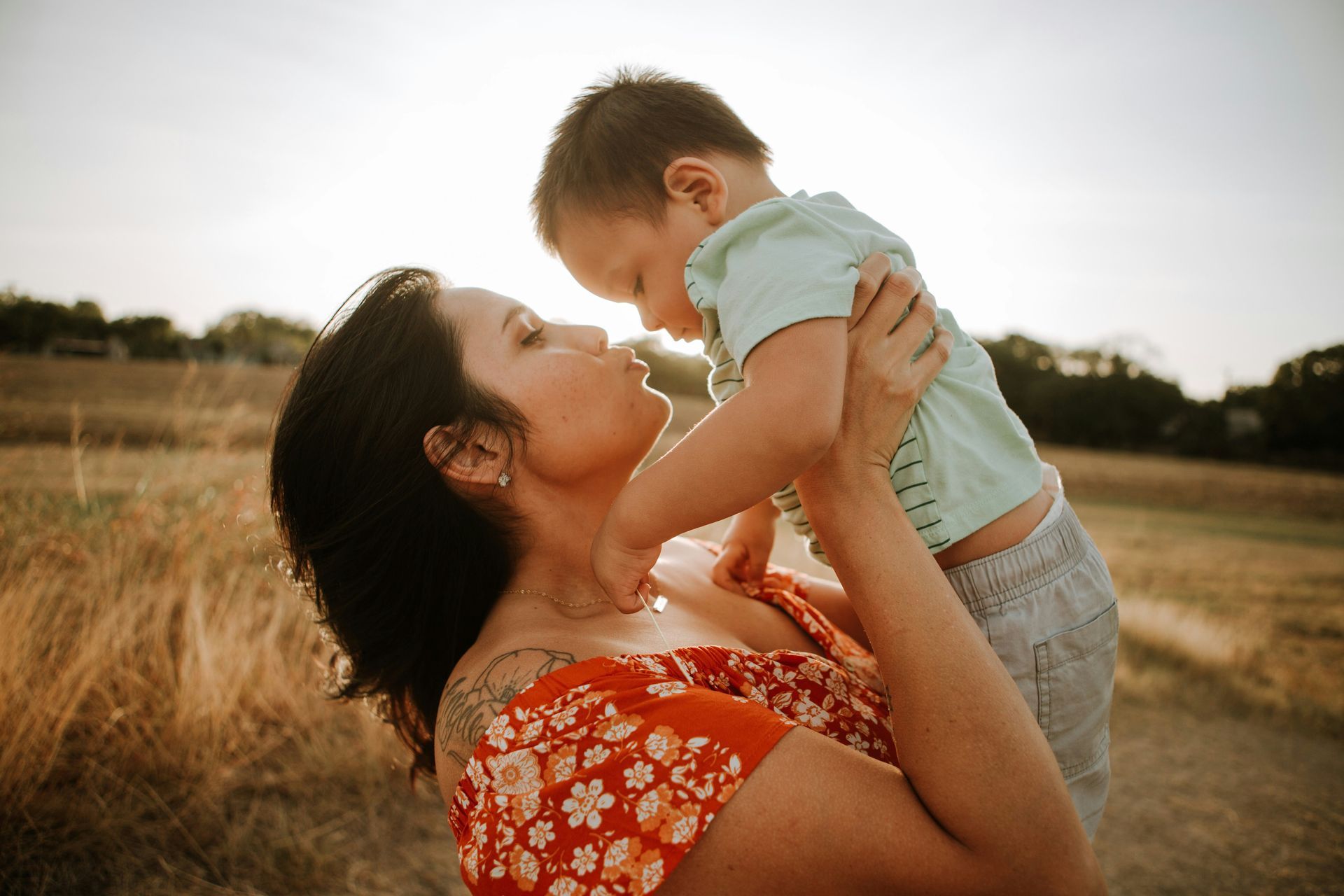 Woman in red dress holds up a child in the sunlight, touching noses in a field.