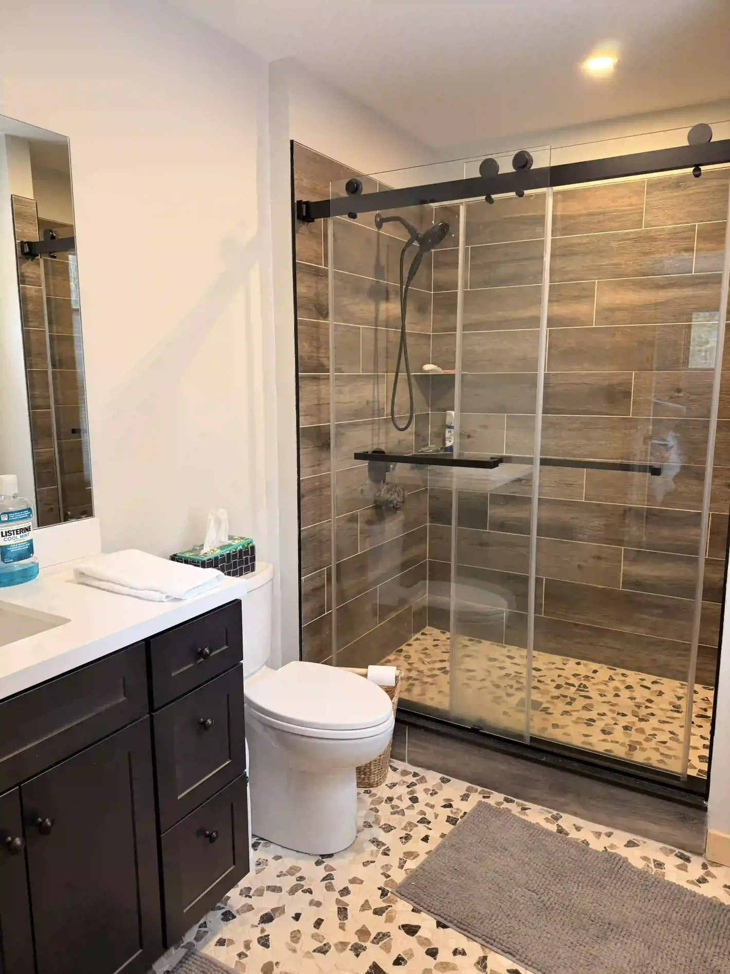Modern bathroom with dark vanity, glass shower enclosure, toilet, and pebble-pattern tile floor.