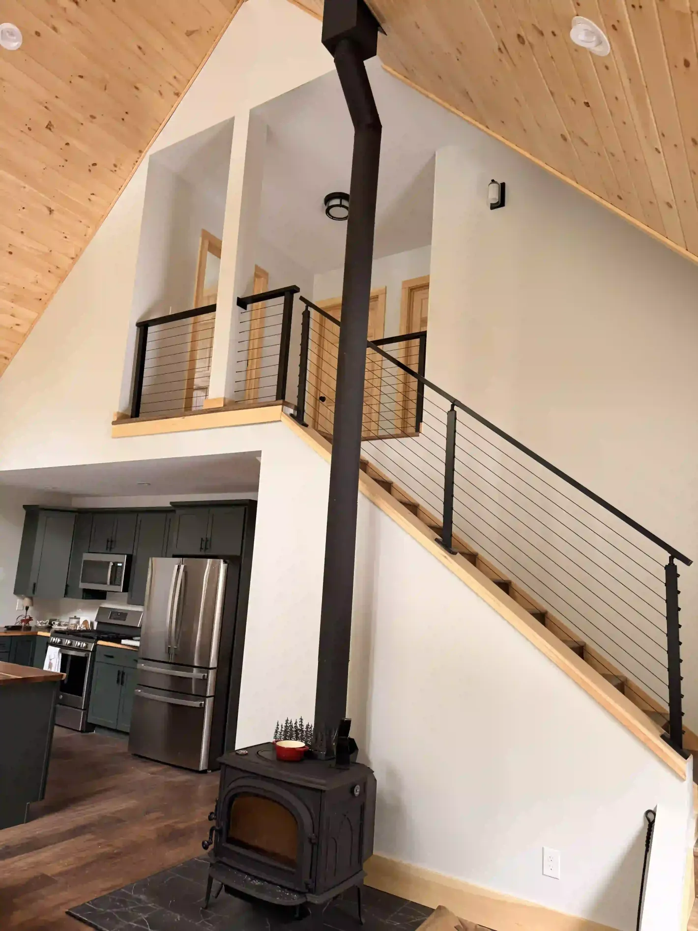 Cozy loft interior with wood stove, staircase, railing, and kitchen below under a sloped wood ceiling