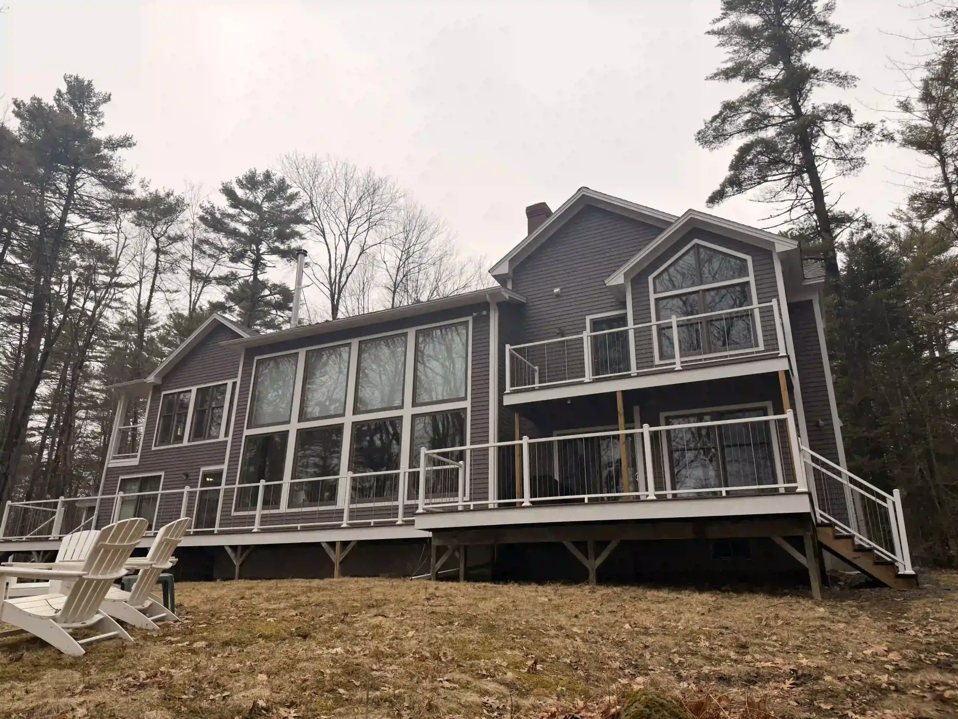 Modern cabin with large windows and deck in a wooded area, with lounge chairs in the foreground