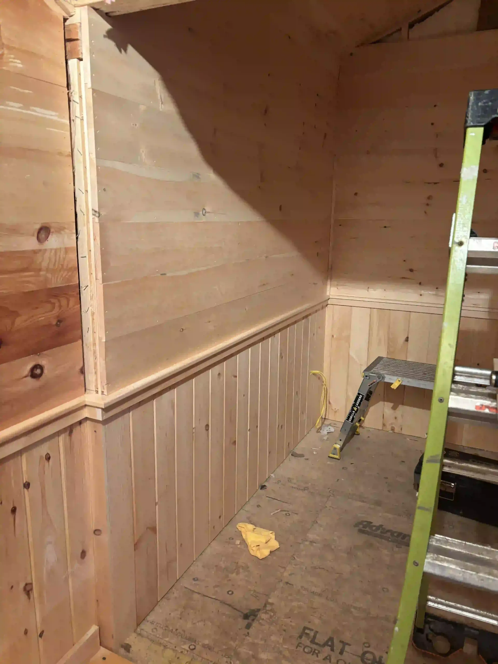 Unfinished wood-paneled room corner with a green ladder leaning against the wall and insulation exposed above.