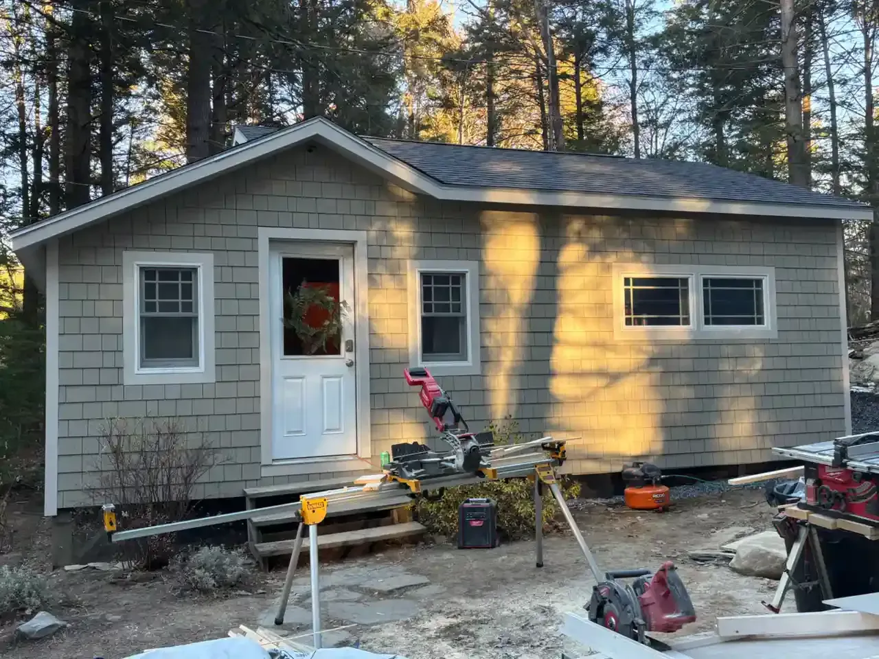 Gray house under construction with tools and materials scattered in front, trees behind.