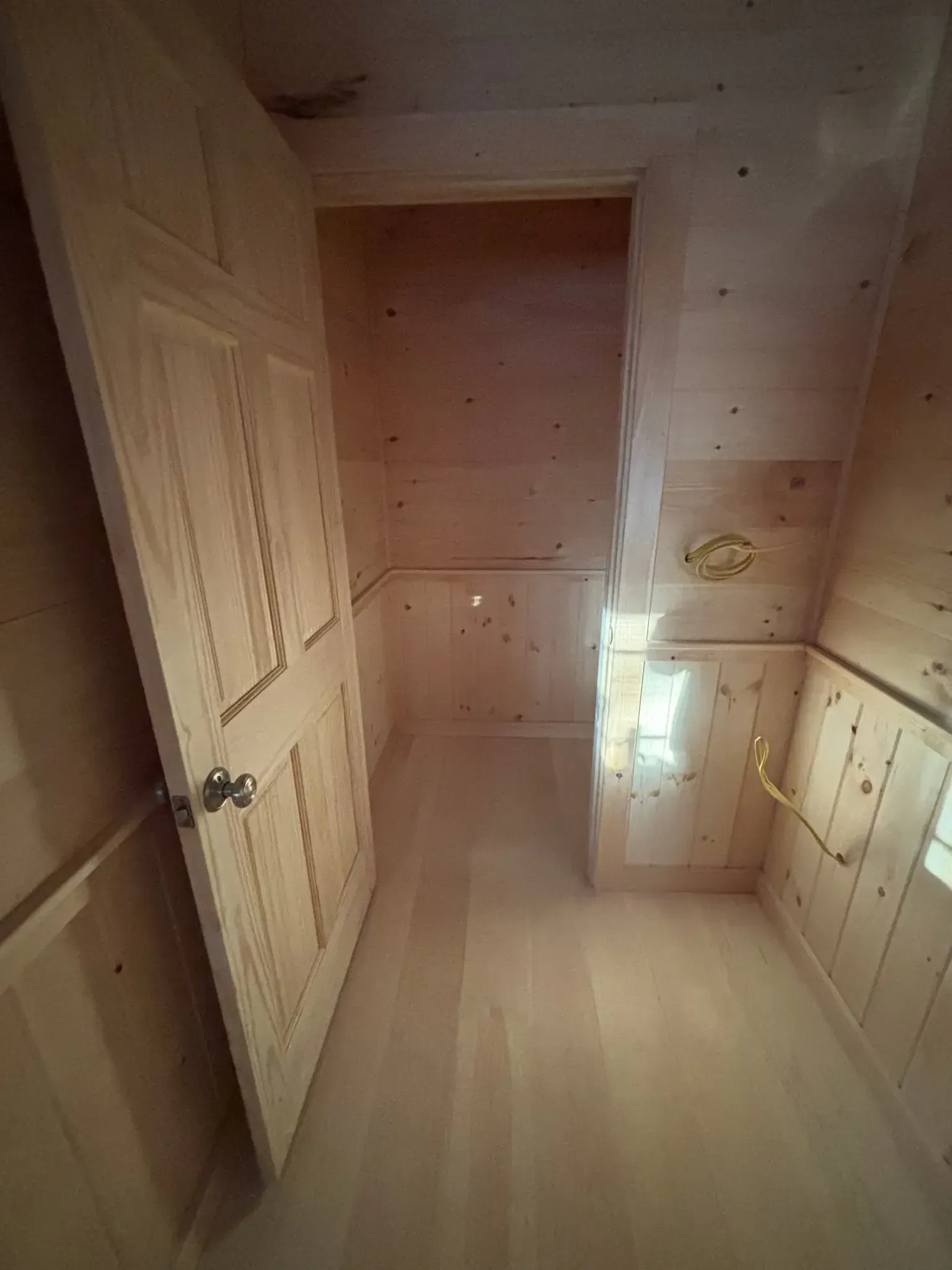 Small unfinished wooden room with built-in cabinets, a door, and light entering from the right.