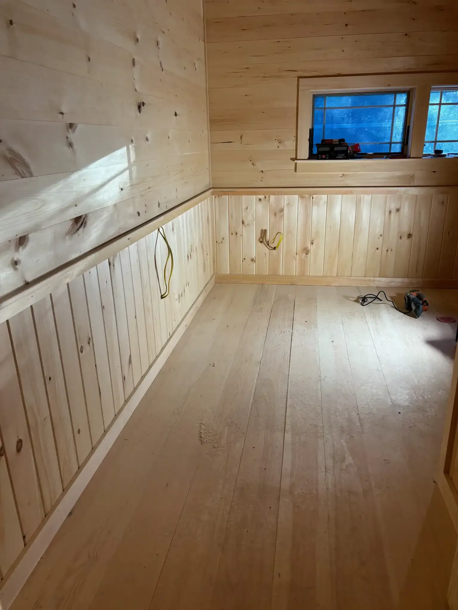 Empty unfinished wooden room with paneled walls, small window, and tools on the floor