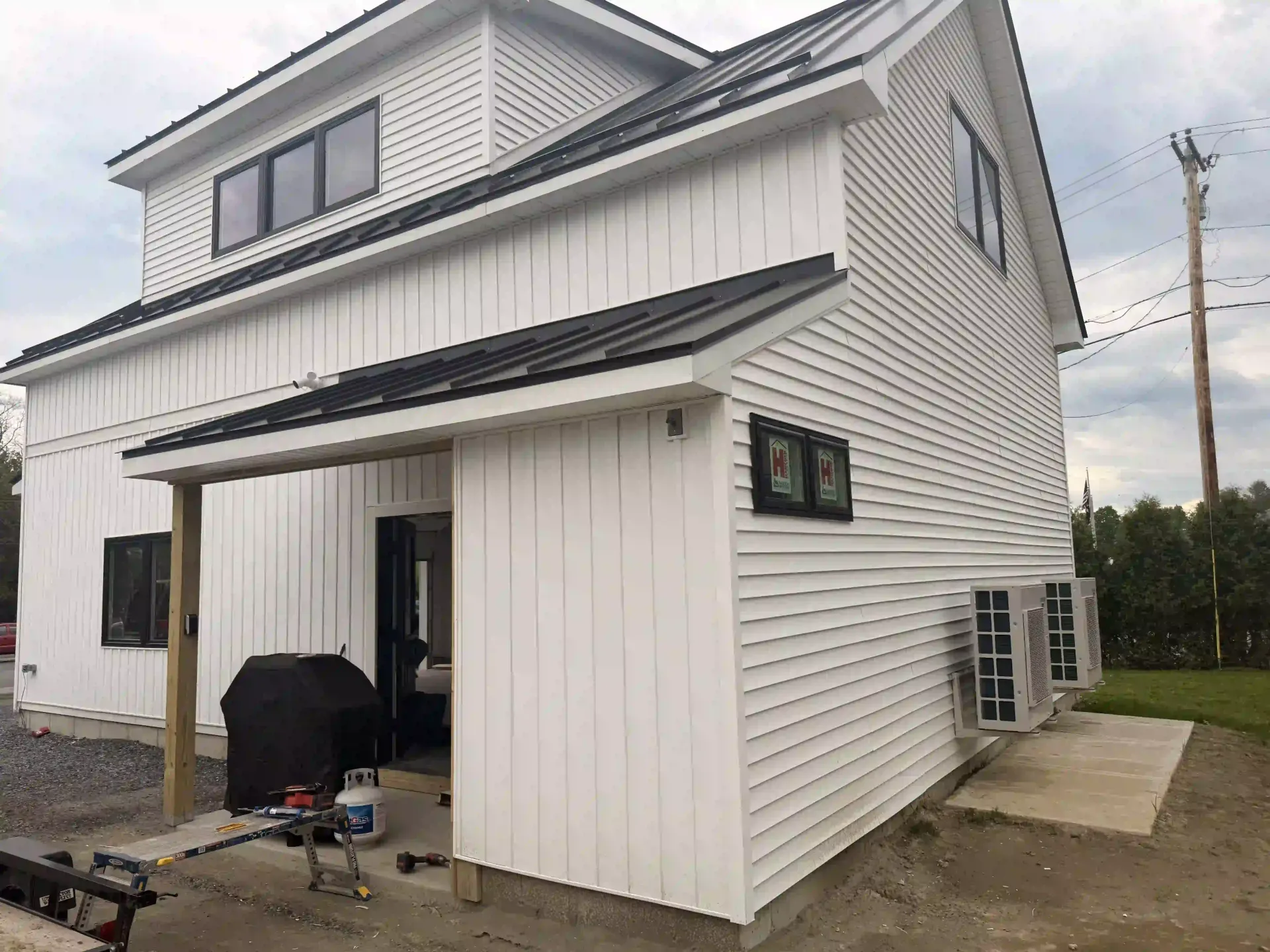 White two-story house with vertical siding, black roof accents, and a small covered porch by a concrete driveway.
