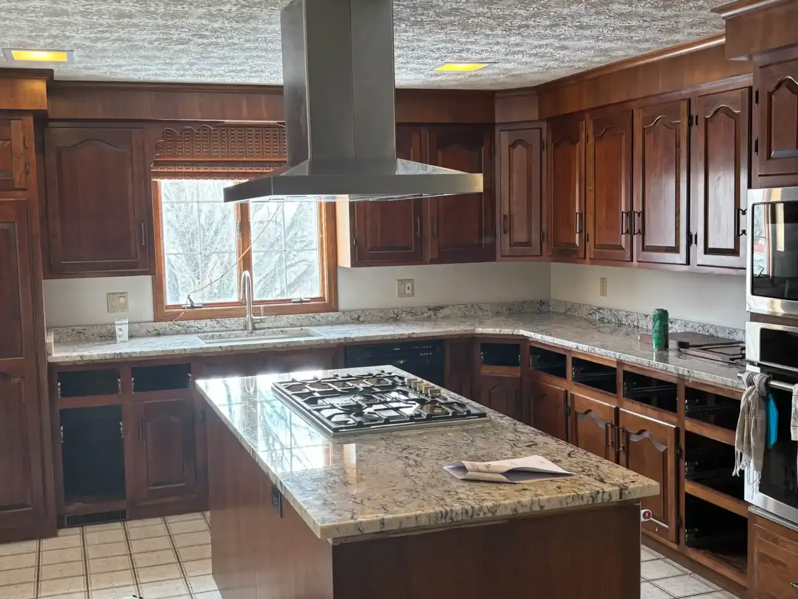 Kitchen with dark wood cabinets, granite island, and stainless range hood by a window.