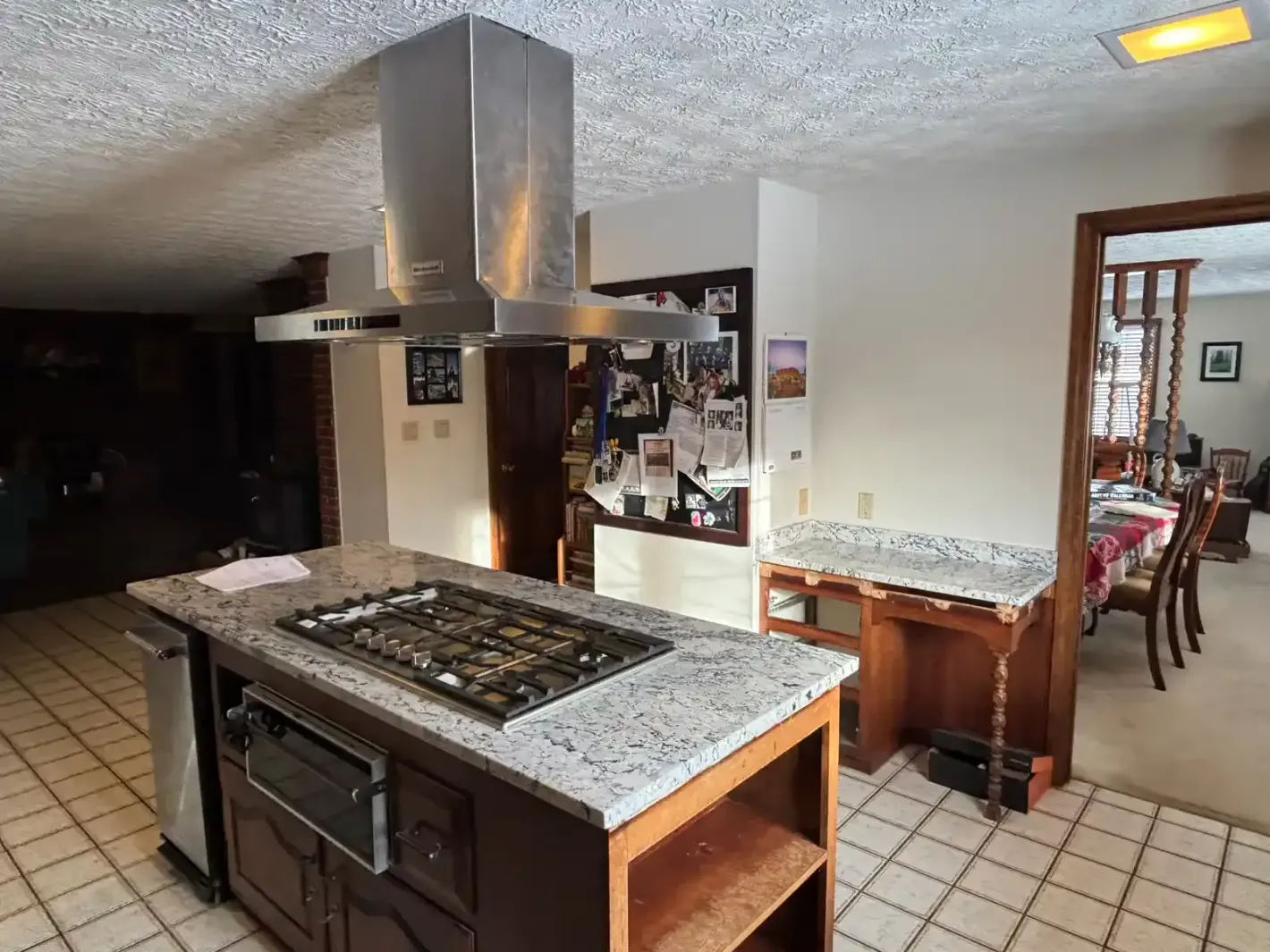 Kitchen with island cooktop, stainless vent hood, granite counters, and tiled floor connected to a dining area
