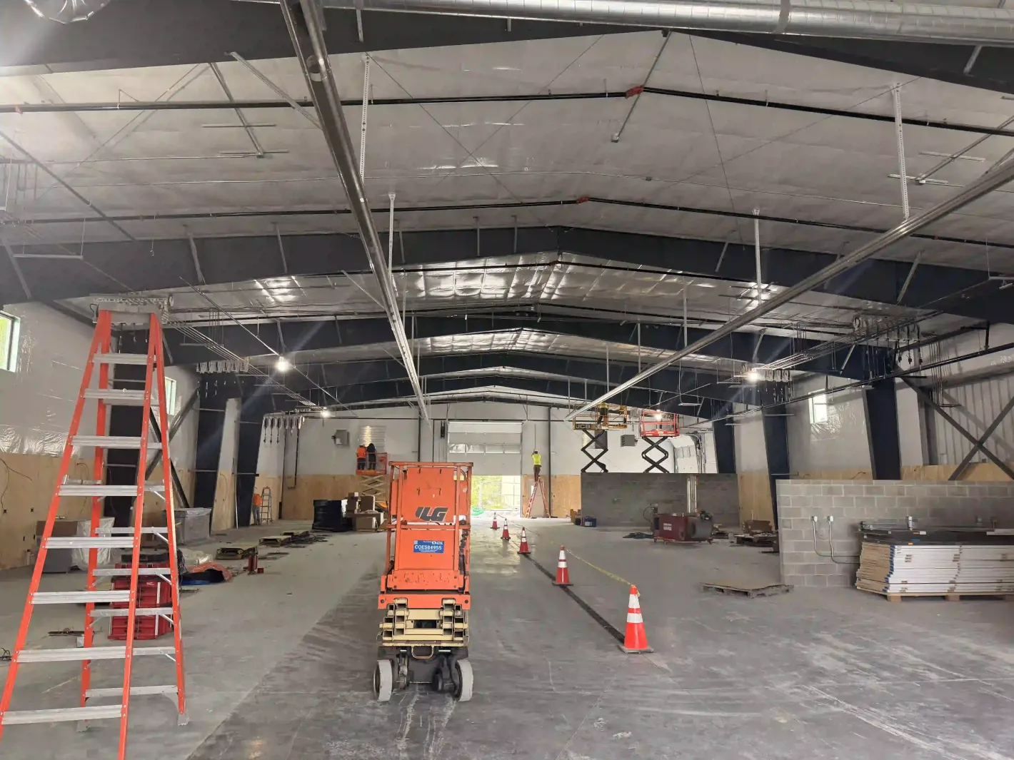 Interior of a warehouse under construction with ladders, a scissor lift, cones, and exposed ceiling framing