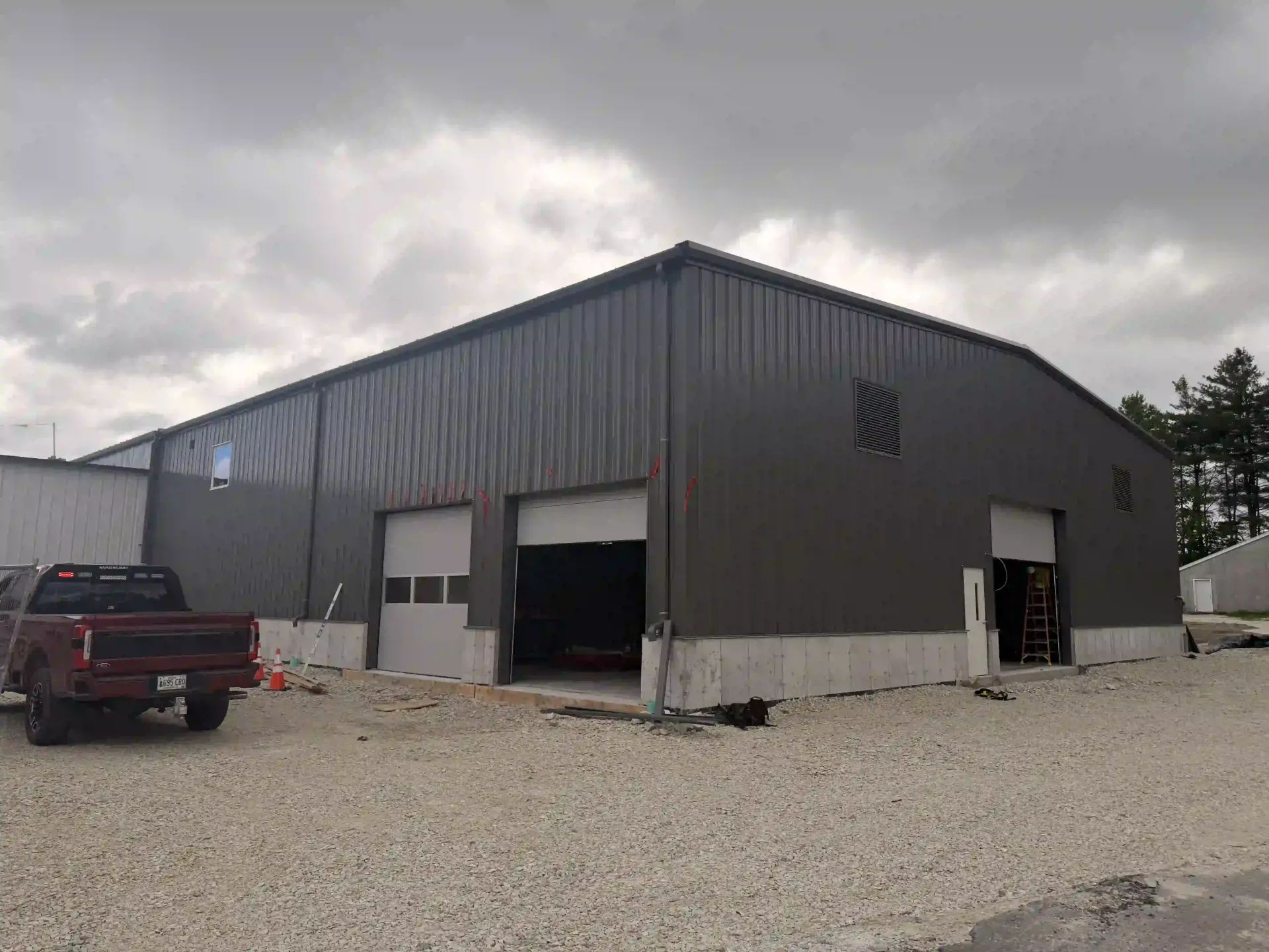 Gray metal workshop with open garage doors and a red pickup parked on a gravel lot under cloudy skies