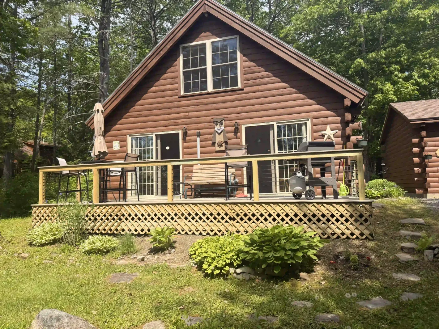 Log cabin with a deck and railing, surrounded by green trees and shrubs.