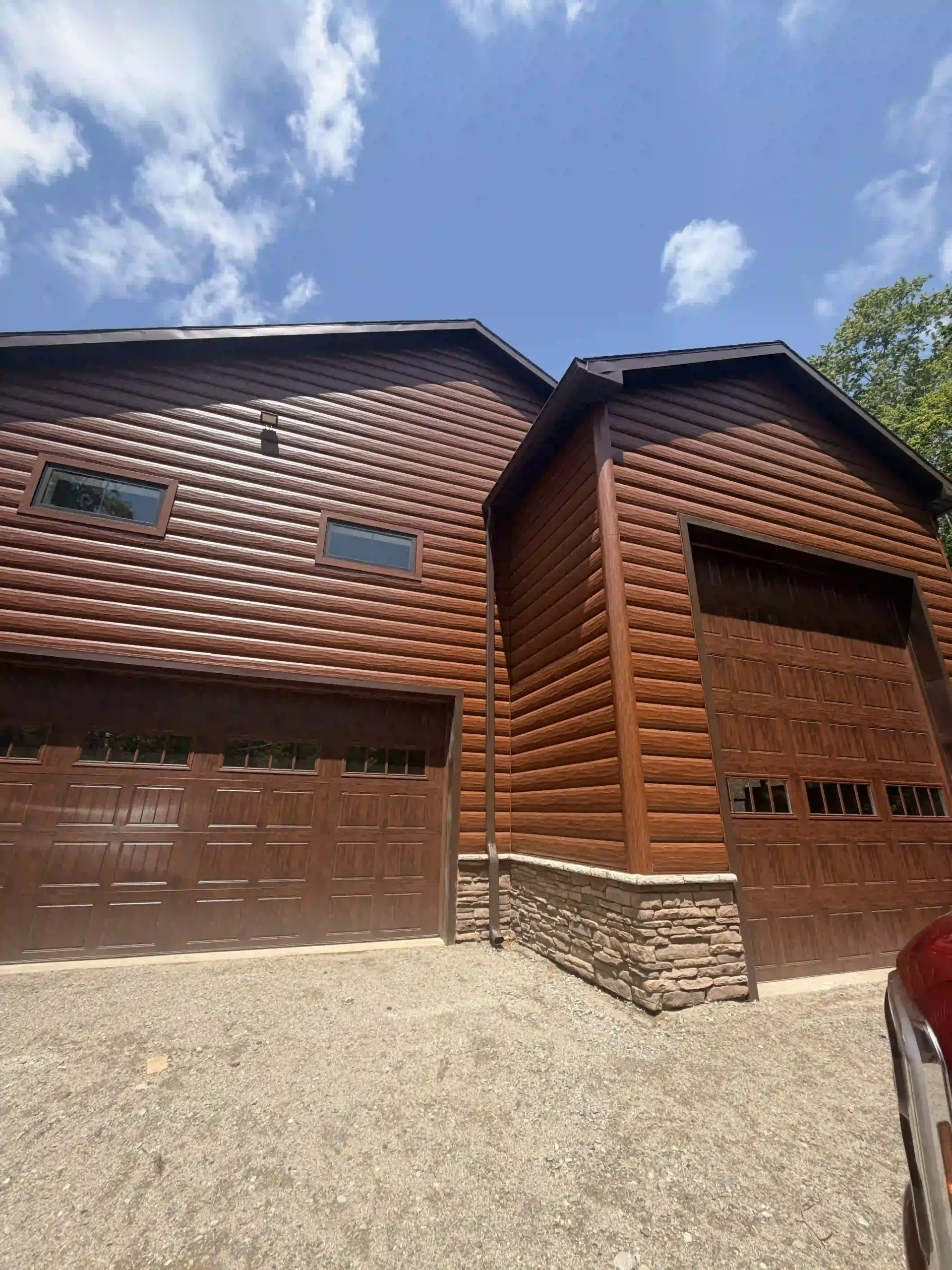 Log cabin-style house with brown garage doors, stone base, and gravel driveway under a blue sky