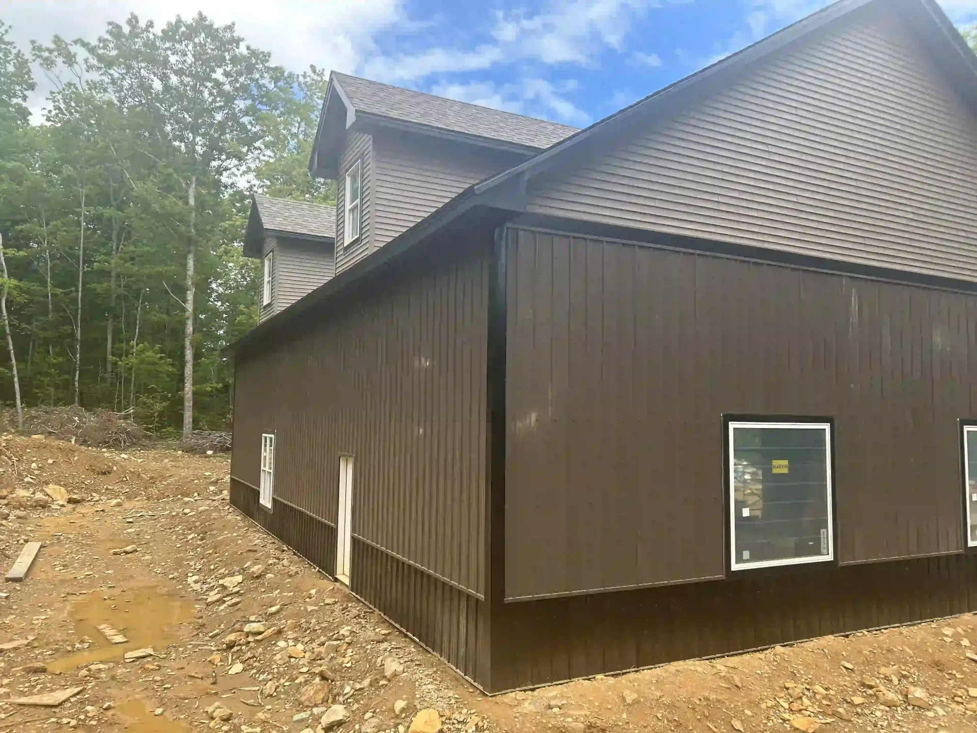 Brown house under construction on a wooded hillside, with bare dirt and a gray roof