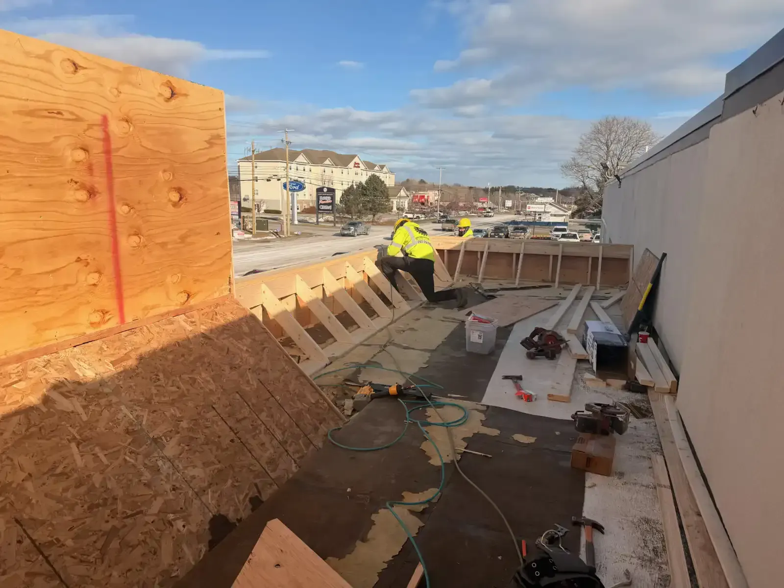 Roofer in yellow jacket working on a flat roof beside plywood walls and tools, with houses in the distance
